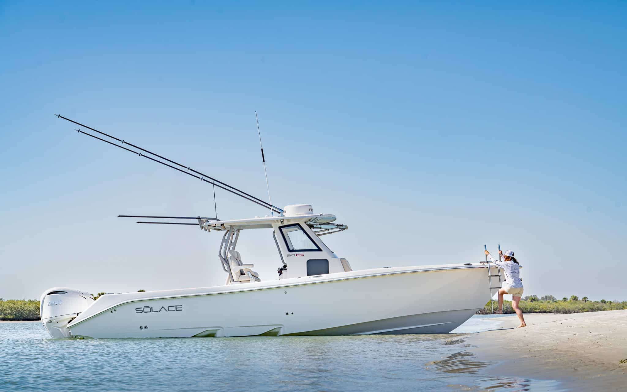 A person steps onto the sand from a sleek Solace boat beached near the shore, fishing rods mounted on the Solace, all under a clear blue sky.