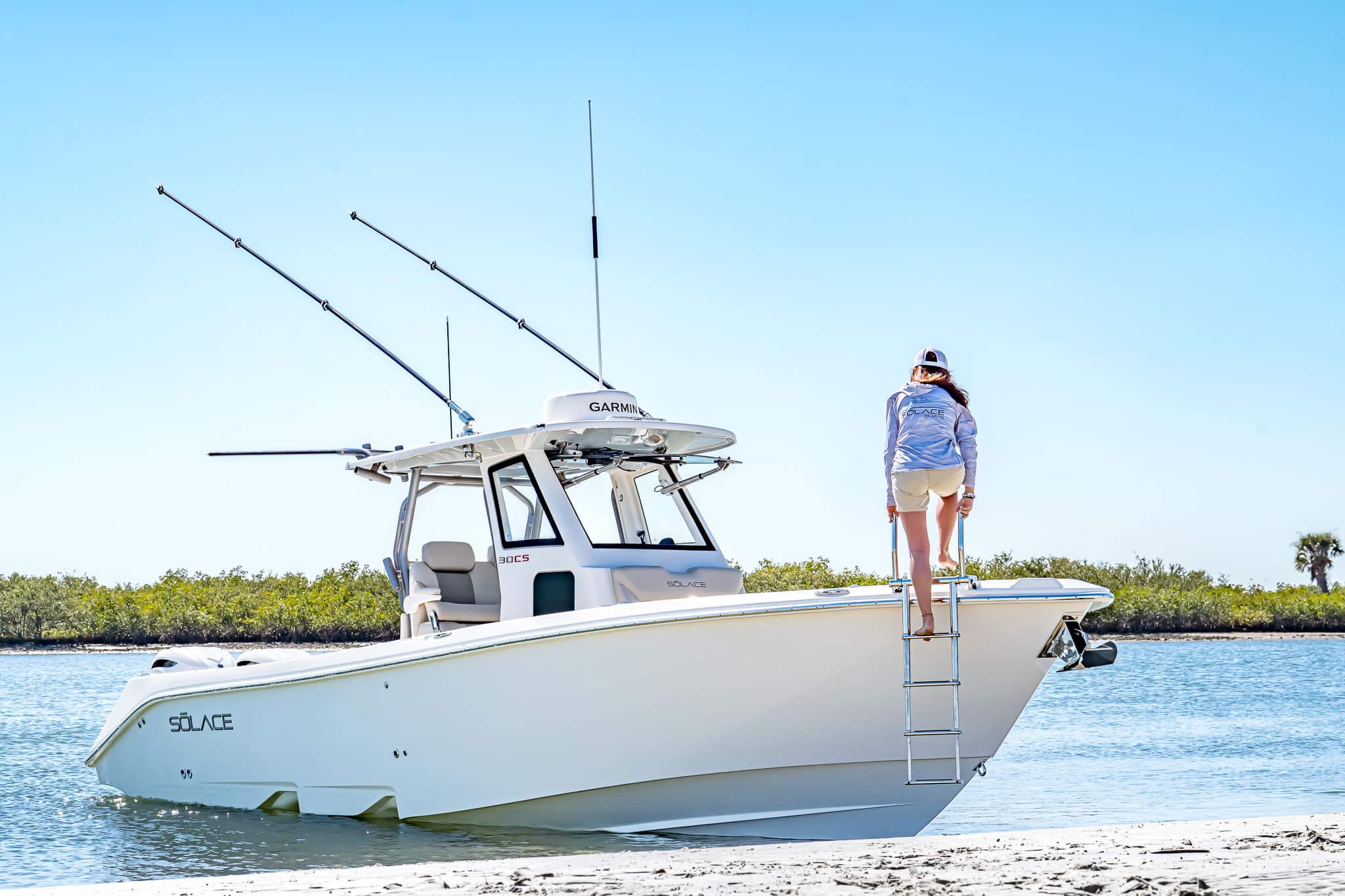 A person climbs a ladder onto a white Solace 30 HCS fishing boat beached near the shore on a sunny day, with fishing rods visible on the boat.