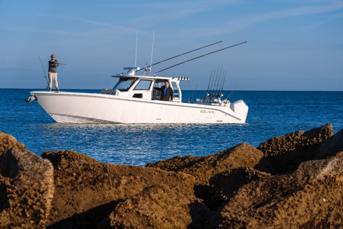 A Solace 41 CS white fishing boat with multiple rods floats near a rocky shore, with one person fishing on the bow and another inside the cabin.